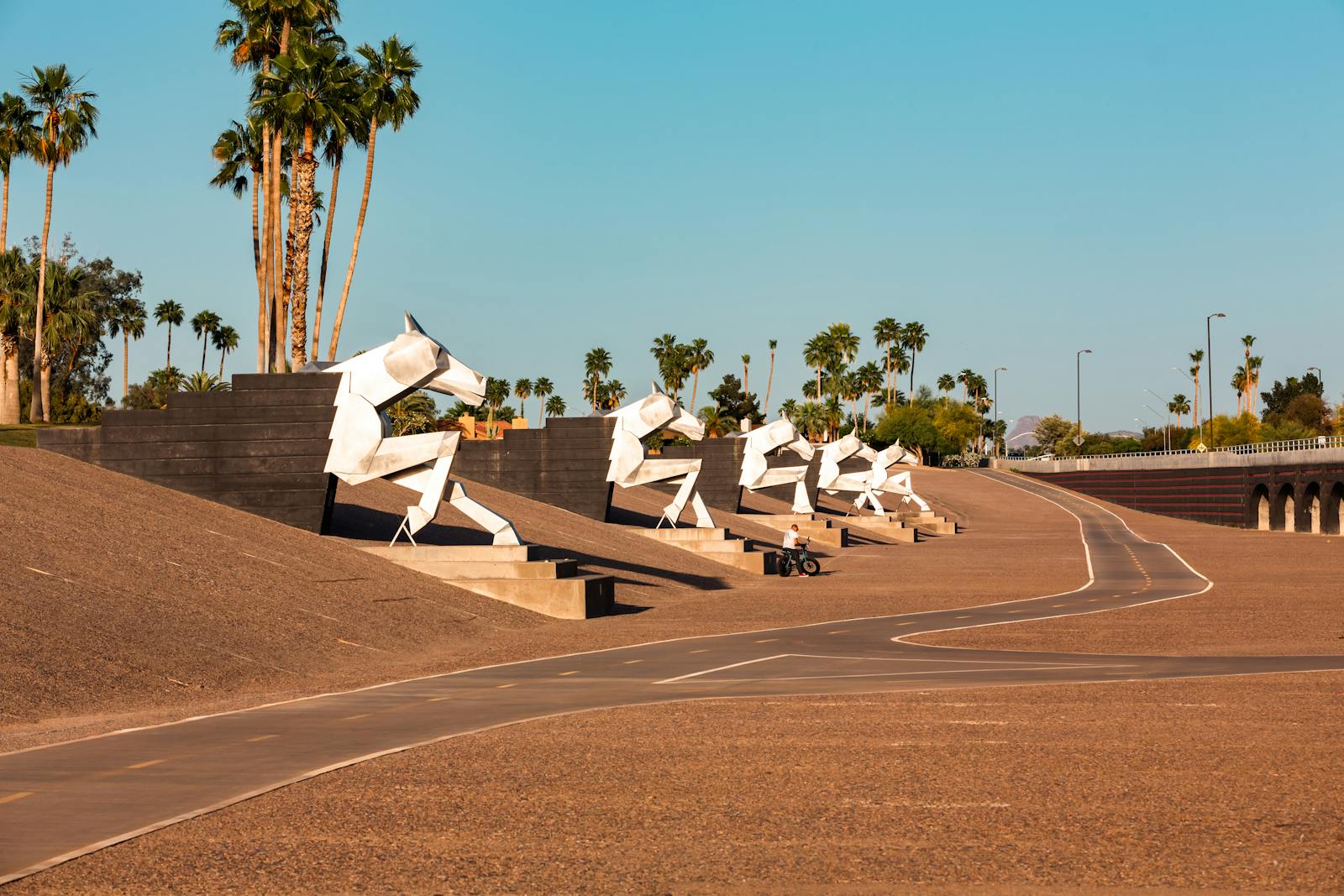 Sculptures of horses line a desert road, surrounded by palm trees and sunny skies.